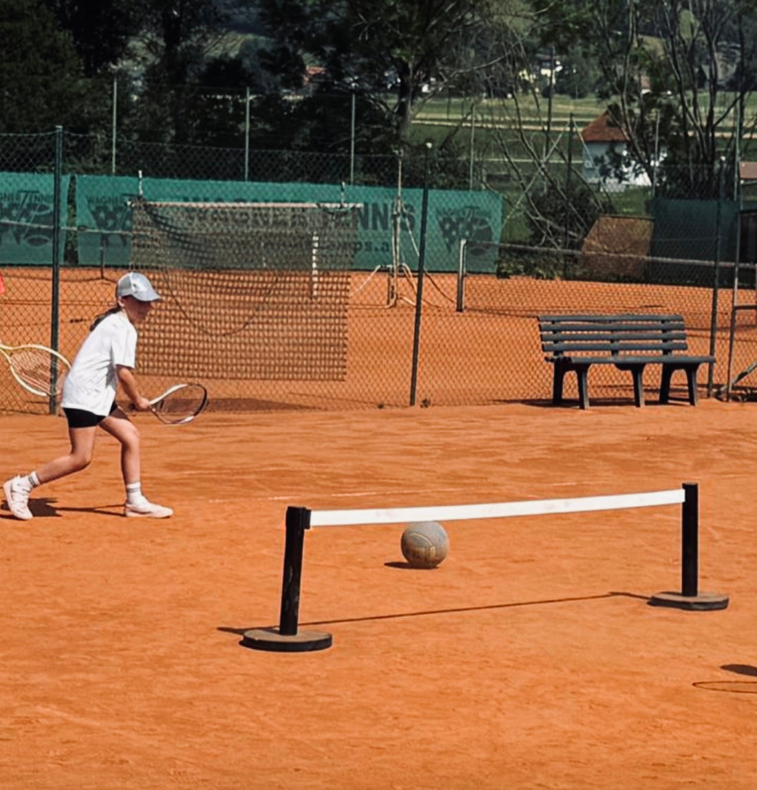 Young children practising tennis with racquets at a low ground-level net on a clay court